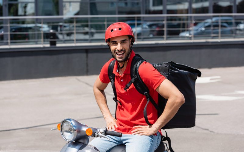 Excited arabian deliveryman with thermo backpack holding hands on hips while sitting on scooter